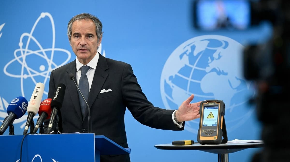 UN nuclear safety official at press conference presenting radiation detection device with IAEA atom logo and world map in background